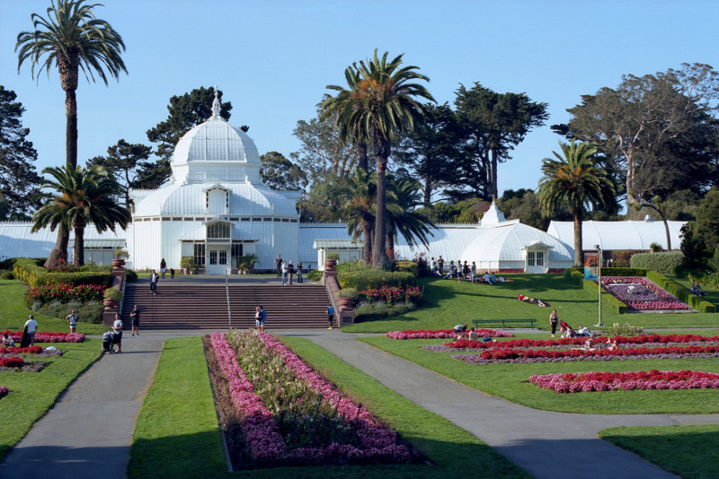 The Hall of Flowers in Golden Gate Park. The building has a central dome with wings extending left and right and perpendicular extensions at each end. It is all panes of glass, painted white, in a wooden framework.