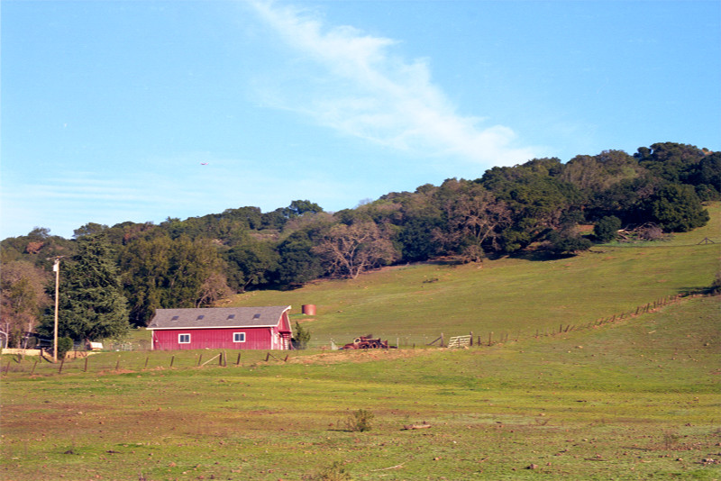 Red barn at lower left, hill with dense forest at crest sloping up toward the right. Little cloudstream sloping up right to left from behind the hill into the blue atmosphere. Near Mckean Road.