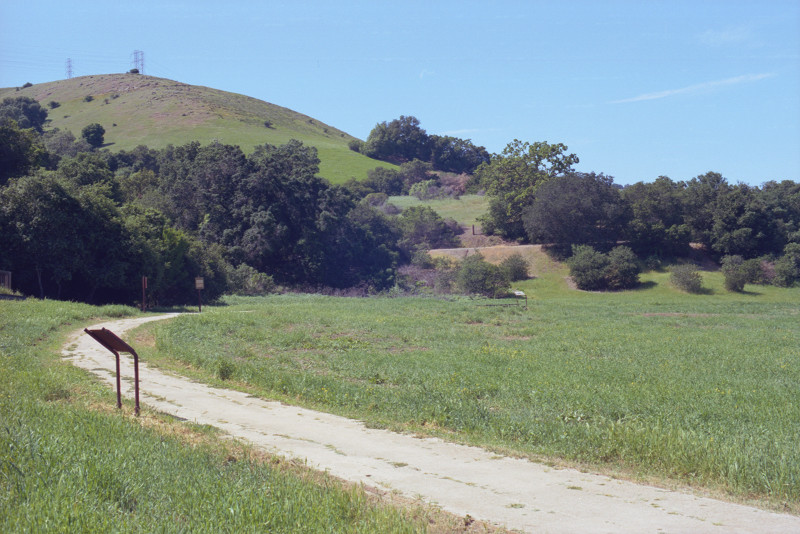 Dusty trail curves from lower right across the photograph to the left and then to the right along the foot of a hill rising up to transmission towers and a clear sky. Bernal Road, near Bernal Ranch. 