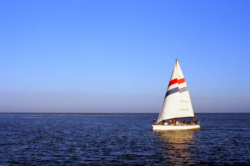 Sloop entering channel to Santa Cruz Yacht Harbor on open sea under open sky. Viewed from Walton Lighthouse at the end of the lighthouse jetty.