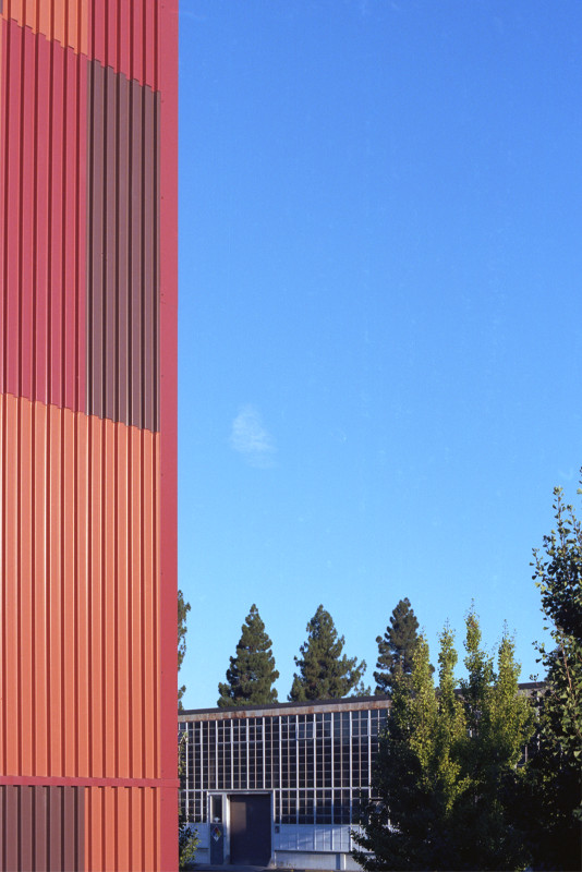 At San José City College, view from a porch with a red slat wall to a building in the distance with evergreens behind it, under a clear blue sky with one tiny cloud floating by.