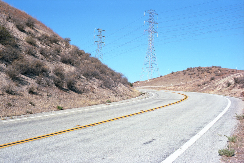 Bailey Avenue climbs and turns to the left around a shrubby hillside. On the right side of the road, centered in the picture, twin transmission towers carrying power for homes and farms.