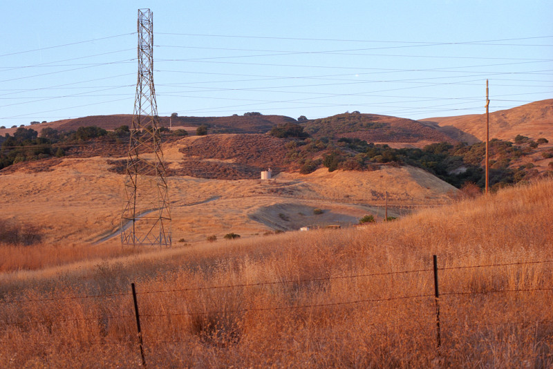 Sunset moments away; view into a valley running along Bailey Avenue, transmission lines running along too. The sere hills turning red in preparation for the display of sunset. Sky still blue.