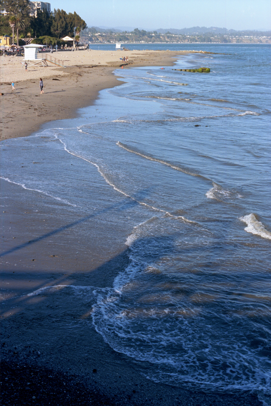 Surf is low at Capitola today, but the curve of the surfline is nice, the white foam edges of the wavelets make patterns over the sand; the shadow of the wharf falls on the beach.