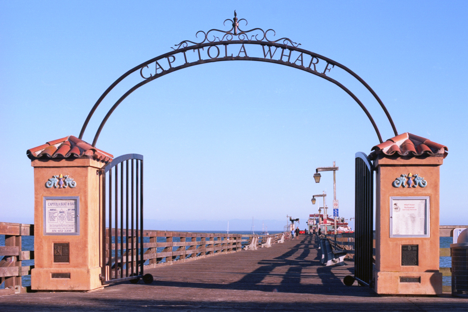 Gate to the Capitola Wharf - rising from two square towers with pointed tile tops, a nice wrought-iron arch over the planks; in the distance, the wooden buildings at the end of the dock. The ocean-sky horizon sea-green below and blue above.