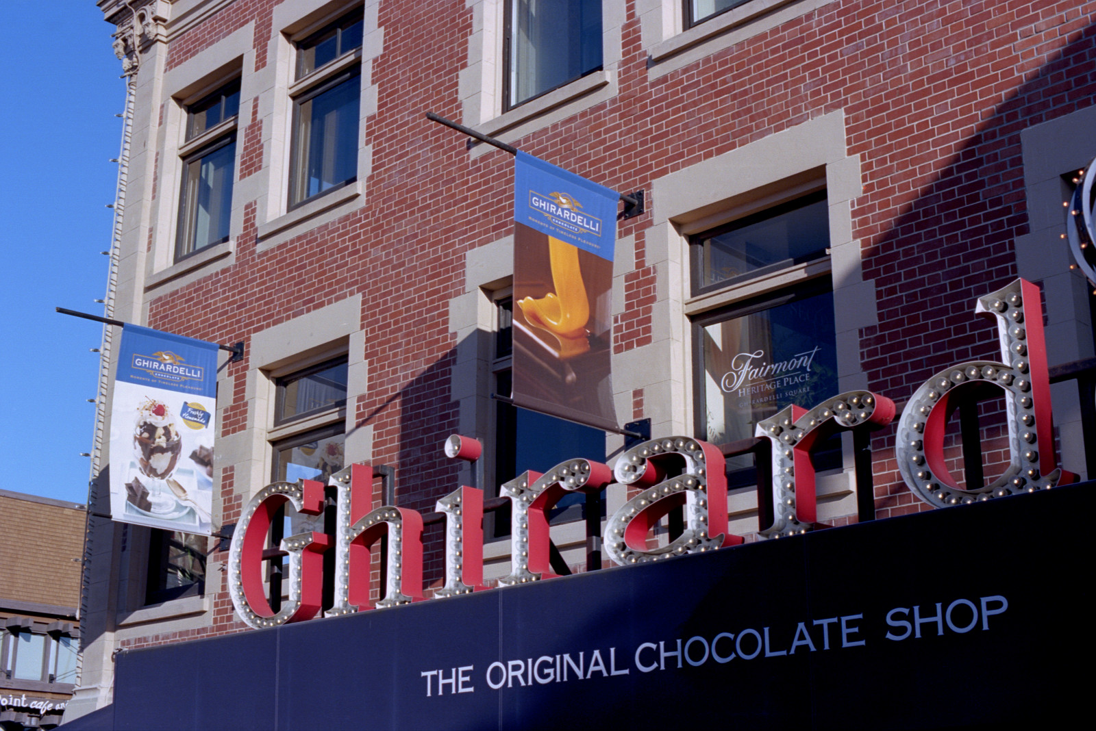 The large red Original Chocolate Shop sign stands above the Chocolate Cafe at Ghirardelli Square. Ghirardelli banners are placed above the marquee, and the background for all is the red brick facade of the building.