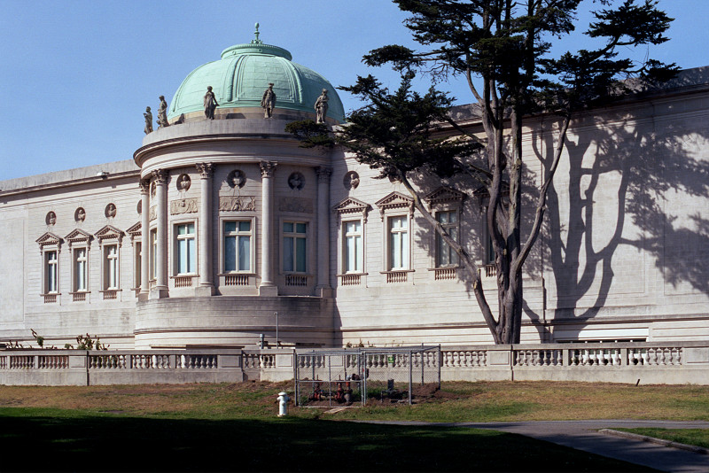 This photo shows the full length of the west elevation of the California Palace of the Legion of Honor, a view not to be missed. A large cypress tree stands next to the Rotunda. There are windows in the wall on both sides of the Rotunda.
