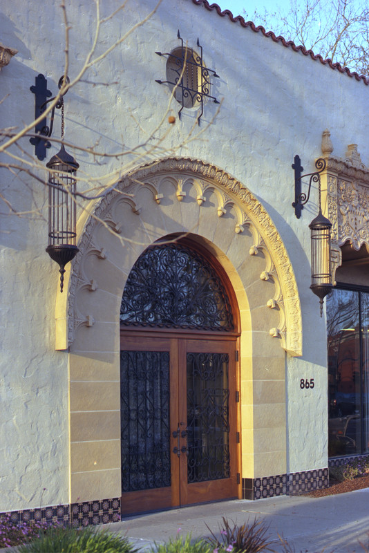 Peach-colored building with arched doorway and window. Wrought iron lantern on both sides of doorway. Small round window with wrought iron grill above the doorway. The Alameda, San José.